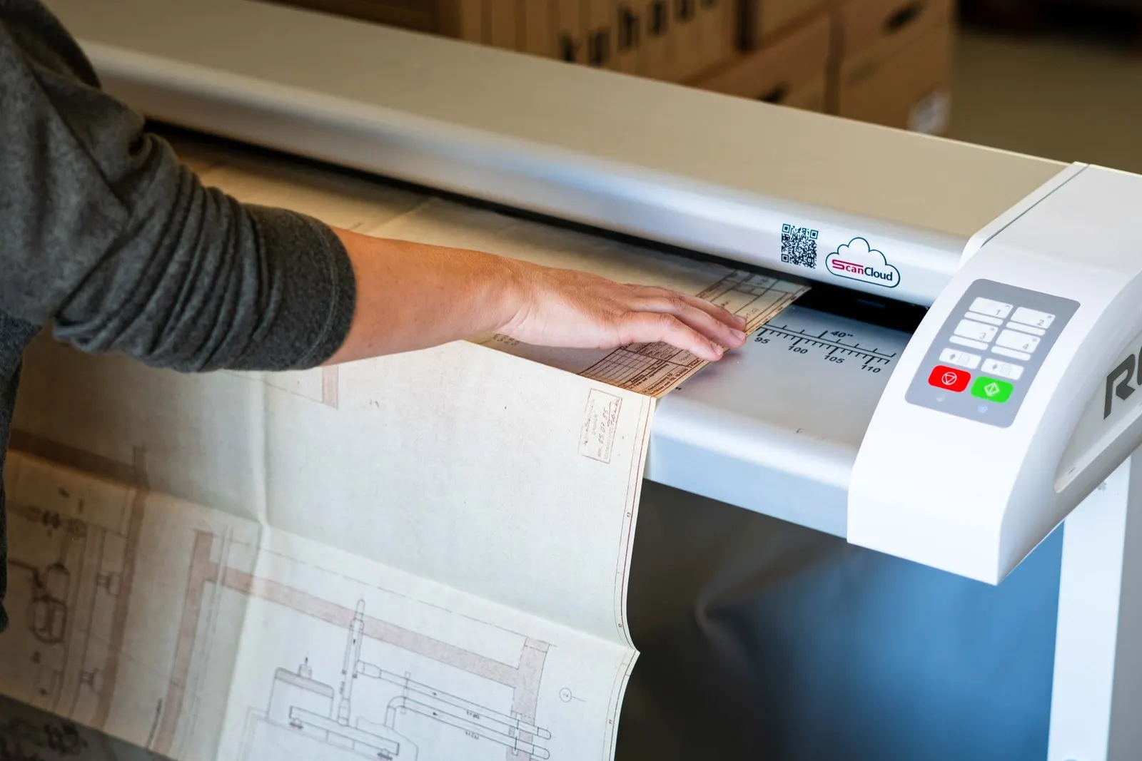 Alter Bauplan wird von Hand in den Großformatscanner eingeführt, Breitenskala bis über 110 Zentimeter sichtbar